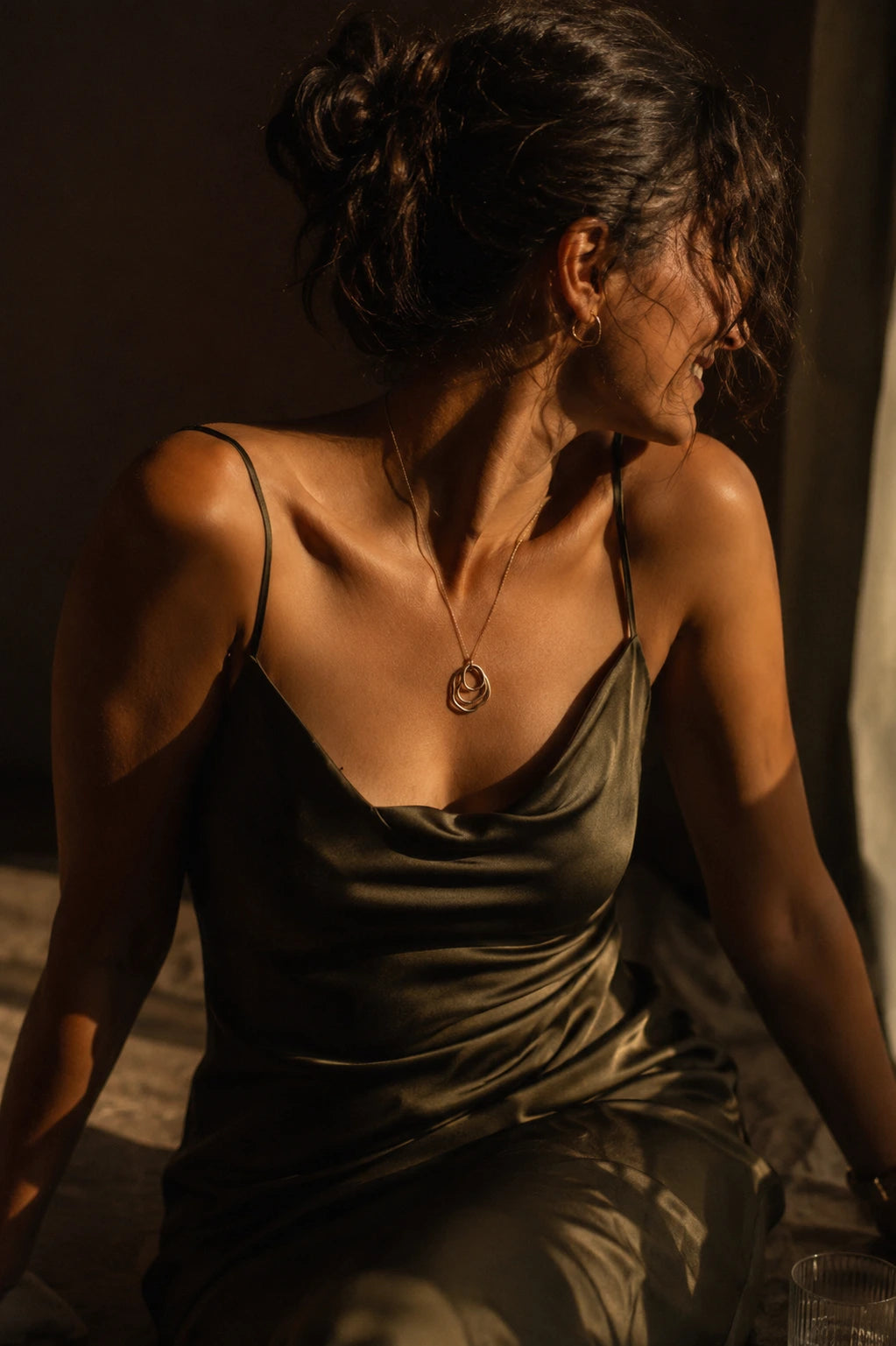 A woman wearing a dark olive green satin spaghetti-strap dress, captured in dramatic warm side lighting against a dark background.