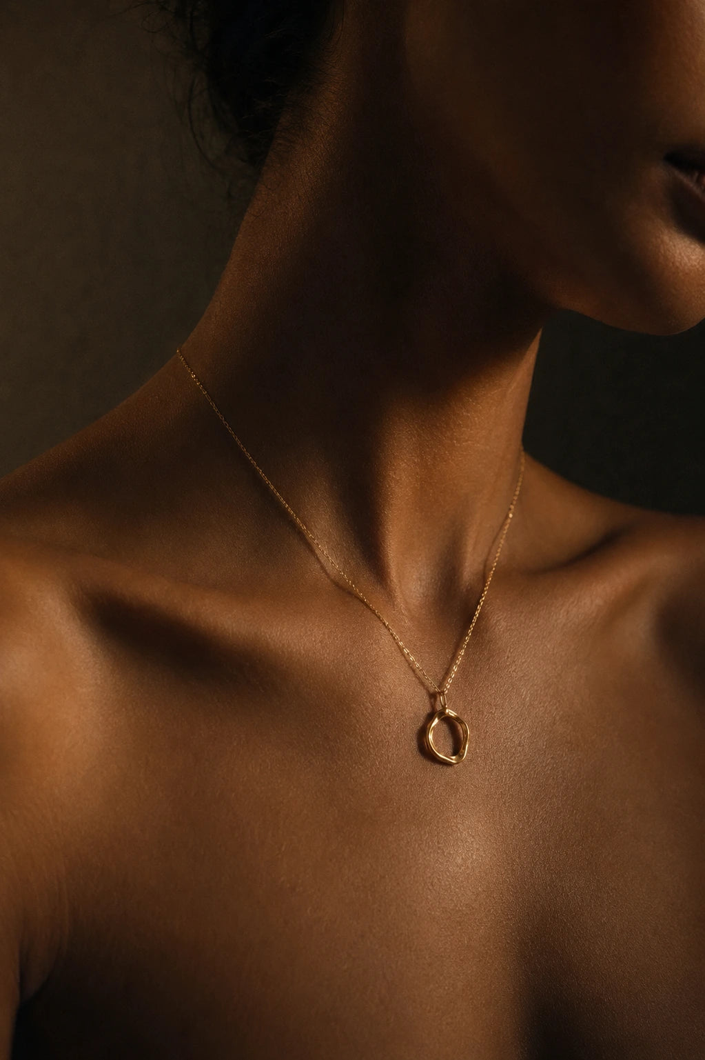 A close-up of a model's neck and upper chest wearing a delicate gold chain necklace with a small open circular pendant, set against a dark, warm-toned background.
